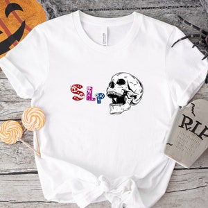 Peut inclure: T-shirt blanc avec un graphique "SLP" coloré et un graphique de crâne noir et blanc. Le crâne est ouvert et a un design détaillé.