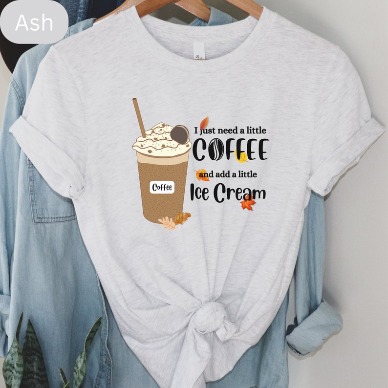 May include: Ash-colored t-shirt featuring a coffee drink graphic with whipped cream, a straw, and the word "Coffee" on the cup. The text "I just need a little coffee and add a little ice cream" is also printed on the shirt. Autumn leaves are included in the design.