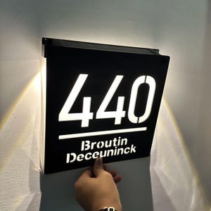 May include: Black rectangular address sign with the number "440" in white, illuminated by a light. The sign also displays the name "Broutin Deceuninck" in white. The sign is mounted on a wall.