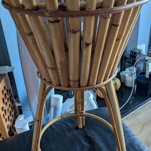 May include: A natural woven bamboo plant stand with a round top and three legs. The stand is made of light brown bamboo.