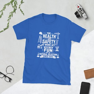 以下が含まれることがあります： ロイヤルブルーの半袖Tシャツ。白のグラフィックテキストで「THE HEALTH AND SAFETY AT WORK ACT TAKING THE FUN OUT OF NATURAL SELECTION SINCE 1974」と書かれ、ヘルメット、はしご、クレーン、建設機械のアイコンが添えられています。