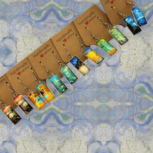May include: A set of eight pairs of silver dangle earrings with rectangular glass charms featuring different paintings. Each earring has a small heart charm with the text "Handmade with love".