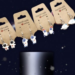 May include: Five pairs of handmade earrings with space-themed charms. The charms include astronauts, rockets, and a spaceship. Each pair of earrings is on a card with the text "Handmade with love".
