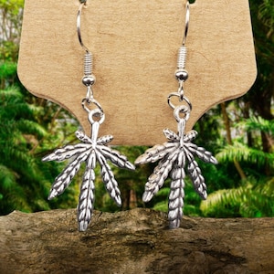 Puede incluir: Pendientes de plata con diseño de hoja de marihuana. Los pendientes cuelgan de una tarjeta marrón.