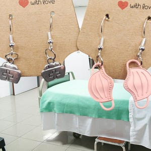 May include: Two pairs of earrings. One pair features a silver charm shaped like a medical symbol, the other pair features a pink charm shaped like a face mask.