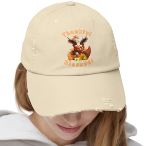 Puede incluir: Gorra de béisbol desgastada de color crema con una vaca de las Tierras Altas de dibujos animados con un tema otoñal. La vaca lleva una bufanda y está sentada en una cornucopia con las palabras "Thankful Blessed!" en naranja.