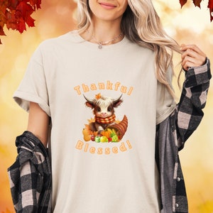 Puede incluir: Camiseta color crema con una vaca de las Tierras Altas de dibujos animados con una bufanda, sentada en una cornucopia con calabazas y calabacines. El texto naranja dice "Thankful Blessed!". El fondo tiene hojas de otoño.