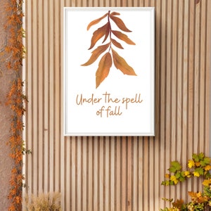 May include: A white framed print with a watercolor illustration of a branch of fall leaves in shades of orange and brown. The text "Under the spell of fall" is written in brown script below the leaves.