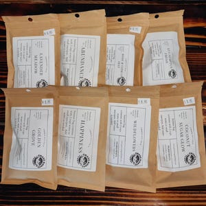 May include: A collection of eight brown paper pouches with white labels, each labeled with a different scent, such as "Lavender Meadow" and "Coconut Sugar Glow." The pouches are arranged on a wooden surface.