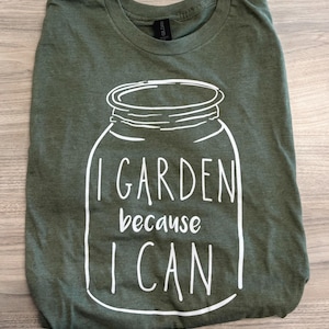 Könnte beinhalten: Olivgrünes T-Shirt mit weißer Grafik eines Einmachglases und dem Text "I GARDEN because I CAN". Dieses weiche T-Shirt ist ideal für Gartenliebhaber und Outdoor-Aktivitäten.