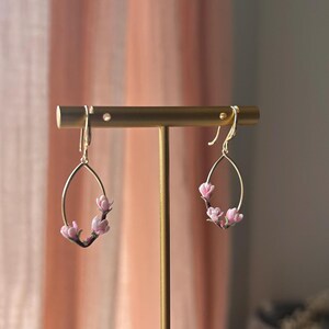 May include: A pair of gold earrings with a delicate oval shape and pink flower accents. The earrings are displayed on a gold stand.