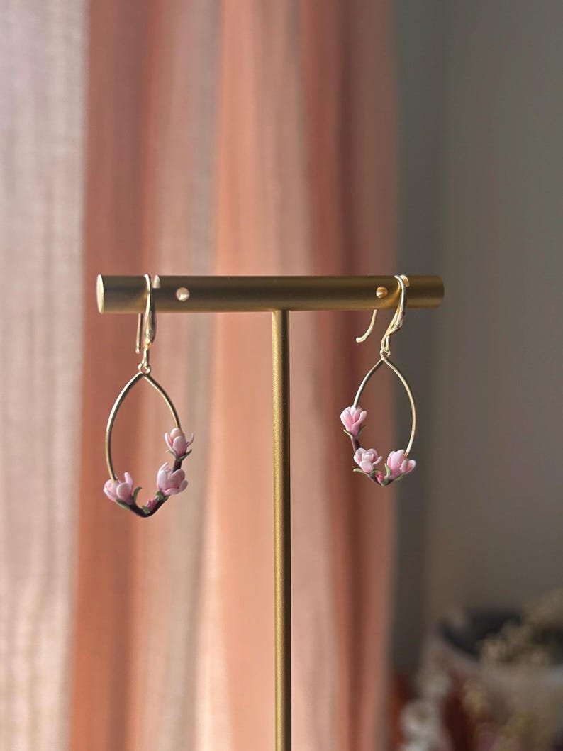 May include: A pair of gold earrings with a delicate oval shape and pink flower accents. The earrings are displayed on a gold stand.