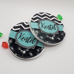 Puede incluir: Dos posavasos redondos de cerámica blanca con un patrón de chevron blanco y negro y una banda azul turquesa. Los posavasos tienen un diseño floral negro y el nombre "Kristel" escrito en cursiva negra.