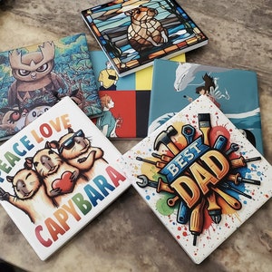 May include: Six colourful ceramic coasters with various designs. One coaster features a cartoon capybara with the text "Peace Love Capybara". Another coaster features a cartoon animal with the text "Best Dad".