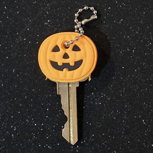 May include: A key with a decorative orange pumpkin-shaped key cover. The pumpkin has a carved face with black eyes, nose, and mouth. A silver ball chain is attached to the top of the key cover.
