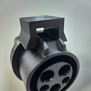 May include: A black electric vehicle charging connector with five circular holes. The connector has a rectangular top with a slot and a rounded base. The image is a close-up shot of the connector.