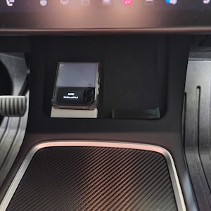 May include: A black smartphone with a clear case is placed in a car's center console. The console has a black carbon fiber-like surface and a white trim.