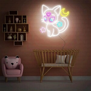 May include: A white neon sign of a cat with a crescent moon and stars. The cat has pink and blue eyes and a pink heart on its forehead. The sign is lit up and is hanging on a wall.