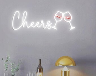 May include: White neon sign with the word "Cheers" and two red wine glasses clinking. The sign is mounted on a grey wall above a wooden table with a gold lamp and wine bottles.