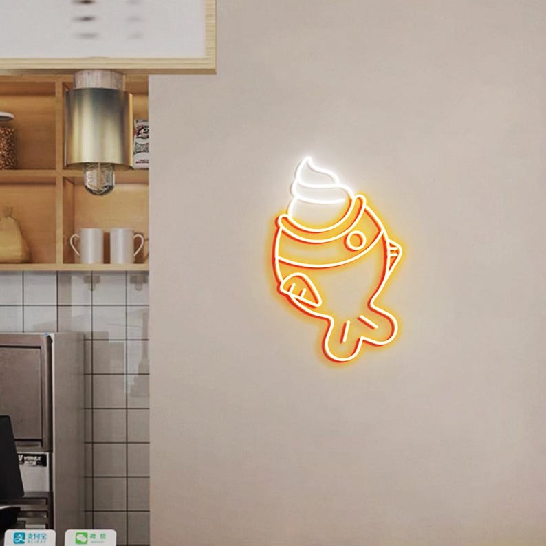 May include: A neon sign in the shape of a fish with ice cream on top. The sign has a bright orange outline with a white ice cream swirl. The sign is mounted on a light beige wall.