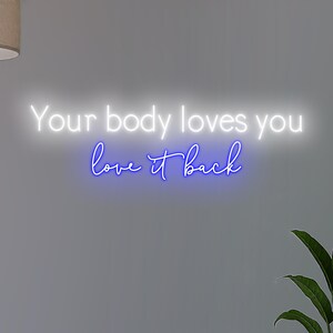 K&ouml;nnte beinhalten: Neonschild mit dem Schriftzug "Your body loves you love it back" in Wei&szlig; und Blau. Das Schild ist an einer grauen Wand angebracht, mit einer Pflanze in der Ecke.