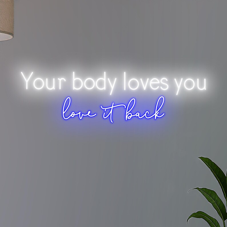 K&ouml;nnte beinhalten: Neonschild mit dem Schriftzug "Your body loves you love it back" in Wei&szlig; und Blau. Das Schild ist an einer grauen Wand angebracht, mit einer Pflanze in der Ecke.