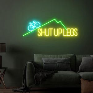 May include: A neon sign with a bicycle and mountain design in green and blue. The words "SHUT UP LEGS" are in yellow. The sign is mounted on a dark green wall above a gray sofa.