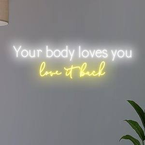 K&ouml;nnte beinhalten: Neonschild mit den Worten "Your body loves you love it back" in Wei&szlig; und Gelb. Das Schild ist an einer grauen Wand angebracht, mit einer Pflanze in der unteren rechten Ecke.