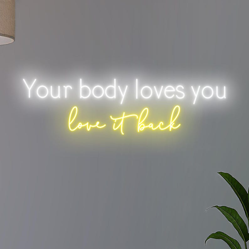 K&ouml;nnte beinhalten: Neonschild mit den Worten "Your body loves you love it back" in Wei&szlig; und Gelb. Das Schild ist an einer grauen Wand angebracht, mit einer Pflanze in der unteren rechten Ecke.