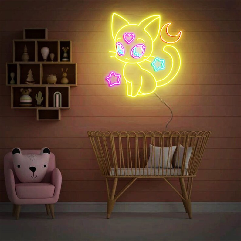 May include: A yellow neon sign of a cat with a crescent moon and stars. The cat has pink and blue eyes and a pink heart on its forehead. The sign is hanging on a pink wall above a crib.