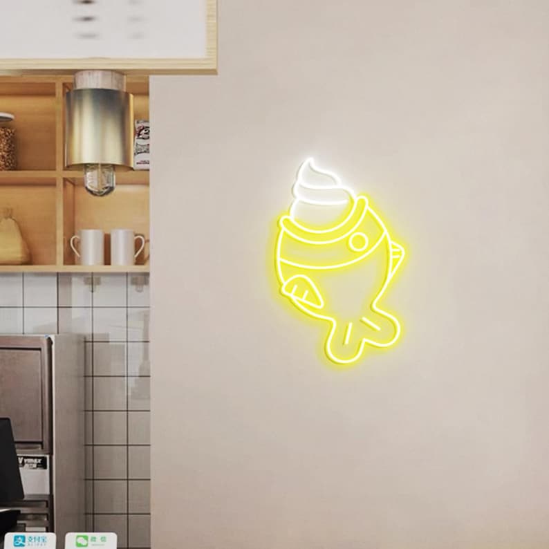 May include: A neon sign in the shape of a fish with a swirl of white cream on top. The sign is yellow and white and is mounted on a beige wall. The sign is likely for a dessert shop or cafe.