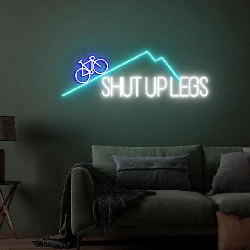 May include: A neon sign with a blue bicycle graphic and the words "SHUT UP LEGS" in white. The sign also features a turquoise mountain range design. The sign is mounted on a dark green wall above a green sofa.