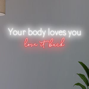 K&ouml;nnte beinhalten: Neonschild mit dem Text "Your body loves you love it back" in Wei&szlig; und Rot. Das Schild ist an einer grauen Wand befestigt, mit einer Pflanze in der unteren rechten Ecke.