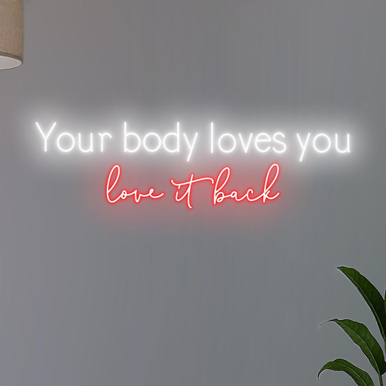 K&ouml;nnte beinhalten: Neonschild mit dem Text "Your body loves you love it back" in Wei&szlig; und Rot. Das Schild ist an einer grauen Wand befestigt, mit einer Pflanze in der unteren rechten Ecke.