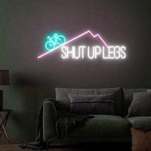 May include: A neon sign with a teal bicycle icon and the words "SHUT UP LEGS" in white. A pink mountain graphic is above the text. The sign is mounted on a dark gray wall above a green sofa.