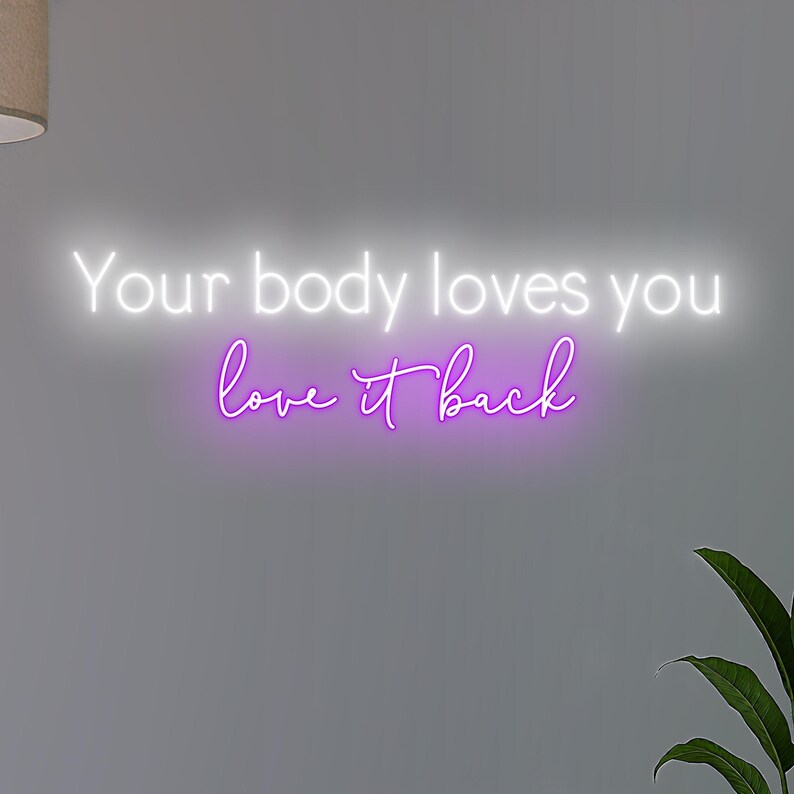 K&ouml;nnte beinhalten: Ein Neonschild mit den Worten "Your body loves you" in Wei&szlig; und "love it back" in Lila. Das Schild ist an einer grauen Wand angebracht, mit einer Pflanze in der unteren rechten Ecke.