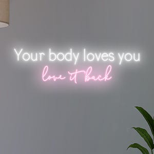 K&ouml;nnte beinhalten: Neonschild mit dem Text "Your body loves you love it back" in wei&szlig; und rosa Schrift. Das Schild ist an einer grauen Wand angebracht, mit einer Pflanze in der unteren rechten Ecke.