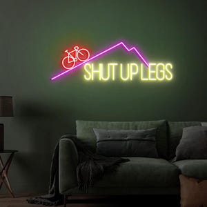 May include: A neon sign with a bicycle icon and the phrase "SHUT UP LEGS" in bright yellow. The bicycle and mountain are in red and purple, respectively. The sign is mounted on a dark green wall above a sofa.