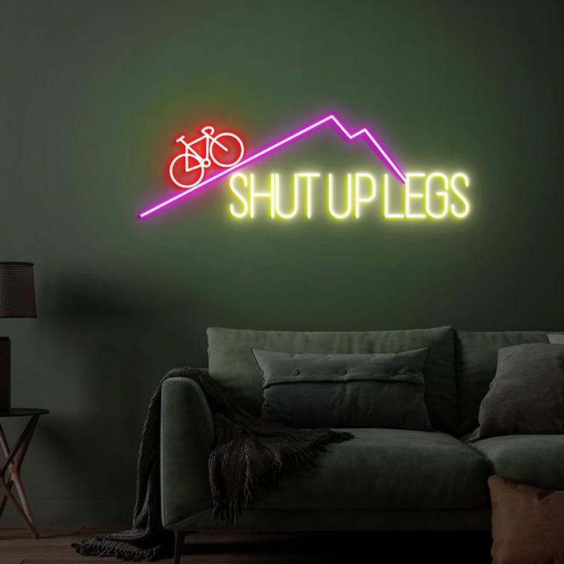May include: A neon sign with a bicycle icon and the phrase "SHUT UP LEGS" in bright yellow. The bicycle and mountain are in red and purple, respectively. The sign is mounted on a dark green wall above a sofa.
