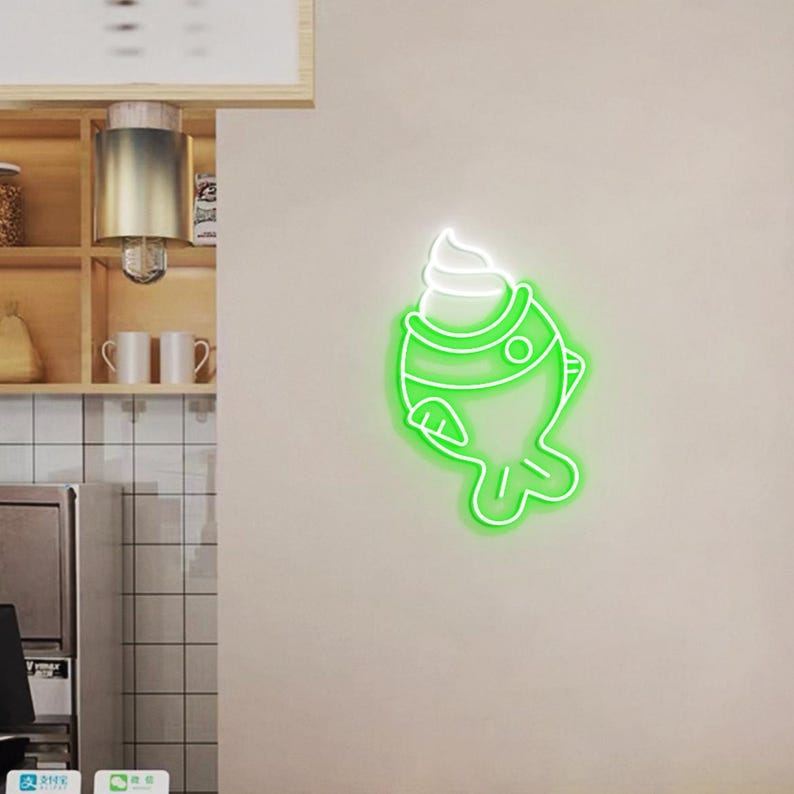 May include: A neon sign in the shape of a fish with a scoop of ice cream on top. The sign is outlined in bright green with a white ice cream topping, mounted on a neutral-colored wall. The sign is a decorative element.