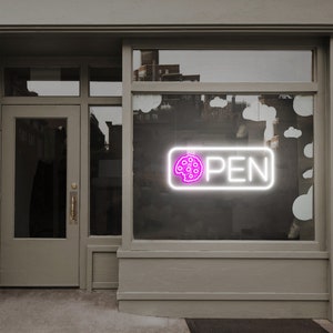 May include: A neon sign in a shop window displays the word "OPEN" in white, with a pink cookie icon. The sign is rectangular with rounded corners. The shop exterior is painted a light gray.