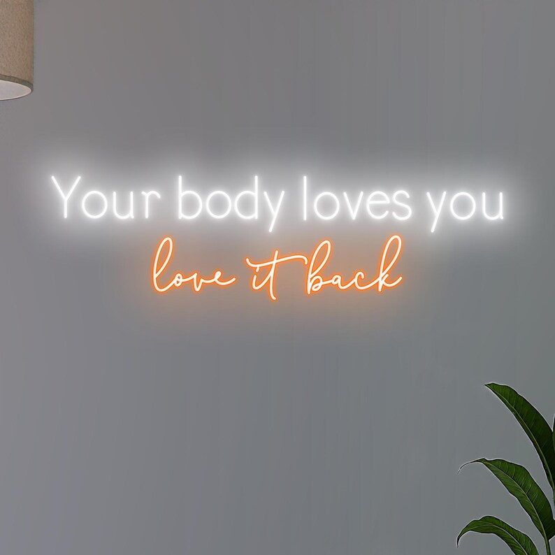 K&ouml;nnte beinhalten: Ein Neonschild mit dem Schriftzug "Your body loves you love it back" in wei&szlig; und orange. Das Schild ist an einer grauen Wand befestigt, mit einer Pflanze in der unteren rechten Ecke.