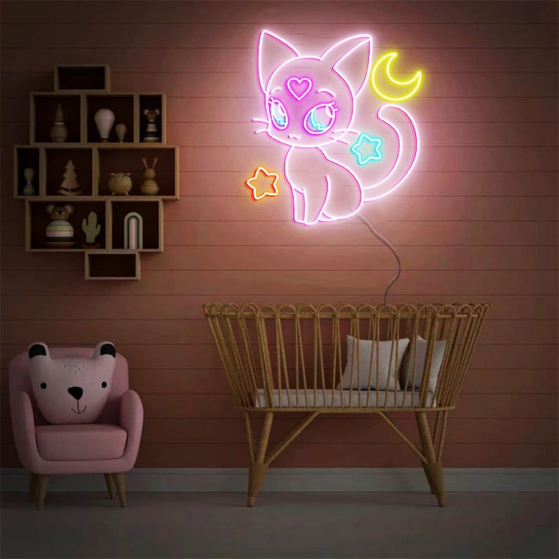 May include: A pink neon sign of a cartoon cat with a heart on its forehead, stars, and a crescent moon. The sign is hanging on a wall with a crib and a pink armchair in the foreground.