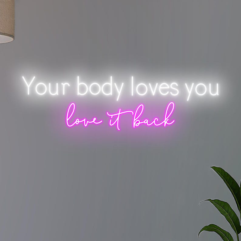 K&ouml;nnte beinhalten: Ein Neonschild mit den Worten "Your body loves you love it back" in Wei&szlig; und Pink. Das Schild ist an einer grauen Wand angebracht, mit einer Pflanze in der unteren rechten Ecke und einer Lampe oben links.