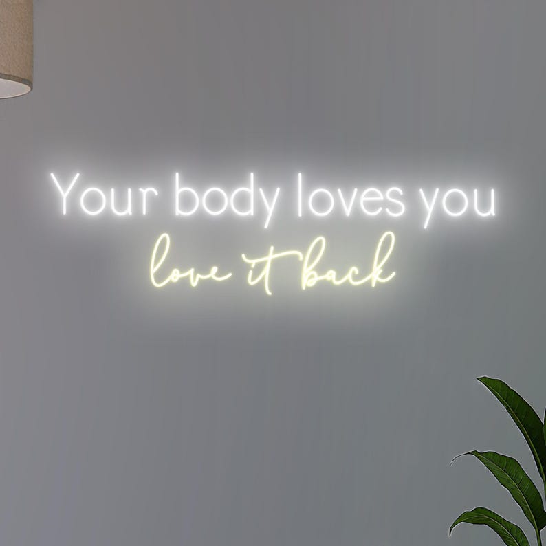 K&ouml;nnte beinhalten: Ein Neonschild mit dem Schriftzug "Your body loves you love it back" in wei&szlig; und gelb. Das Schild ist an einer grauen Wand befestigt, mit einer Pflanze in der unteren rechten Ecke.