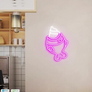 May include: A neon sign in the shape of a fish with a scoop of ice cream on top. The sign is outlined in bright pink with a white ice cream topping. The sign is mounted on a light beige wall.