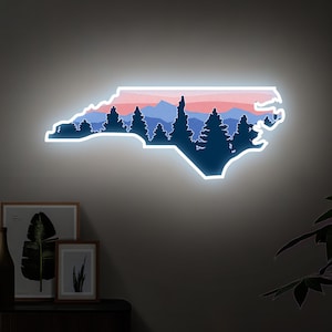May include: A neon sign in the shape of North Carolina, featuring a landscape silhouette. The design includes dark blue trees, mountains, and a gradient sky of pink and blue. The sign is outlined in white and mounted on a gray wall.