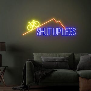 May include: Neon sign with a yellow bicycle, orange mountain range, and the words "SHUT UP LEGS" in blue. The sign is mounted on a dark green wall above a green sofa with pillows.