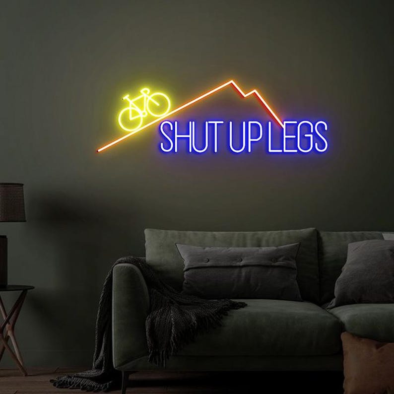 May include: Neon sign with a yellow bicycle, orange mountain range, and the words "SHUT UP LEGS" in blue. The sign is mounted on a dark green wall above a green sofa with pillows.