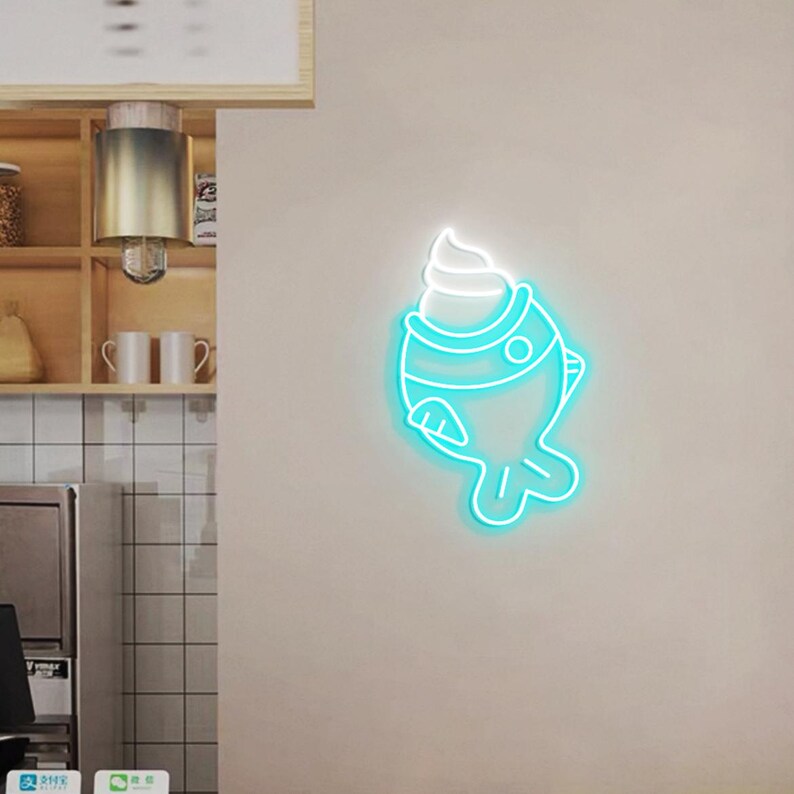 May include: A teal neon sign in the shape of a fish with a swirl of white cream on top. The sign is mounted on a light beige wall. The fish design has a round eye and a fin.